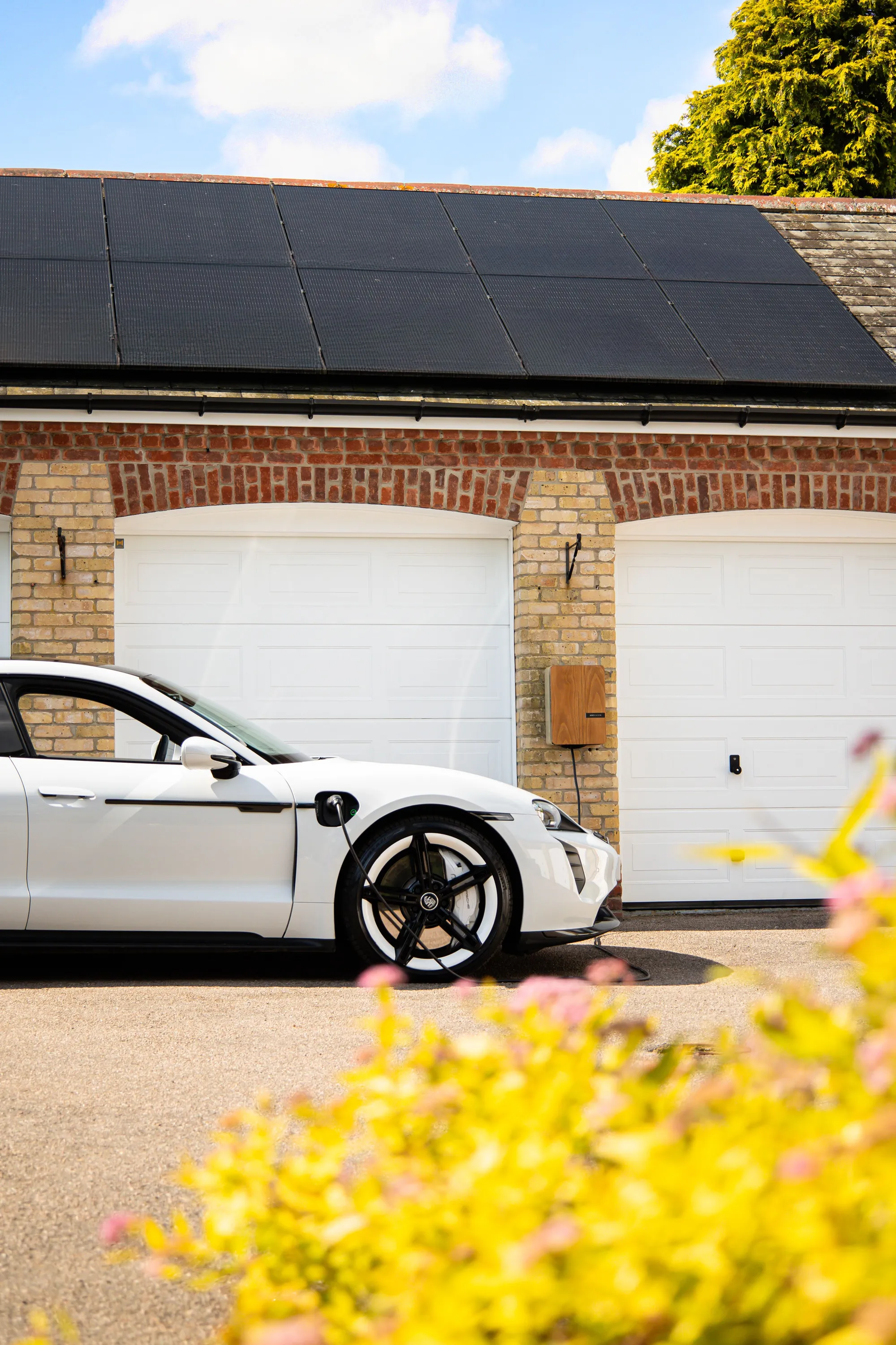 Electric car charging from a wall-mounted home charger beside garages with a solar roof